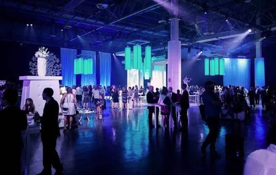 Attendees networking in a blue-lit expo hall with lounge areas and installations during an evening reception