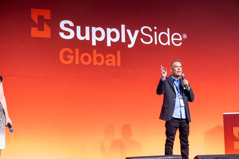 A speaker presenting on stage in front of the SupplySide Global logo screen